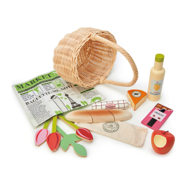 A hand crafted wicker basket and newspaper, bar of fake chocolate, bottle of lemonade, continental salami, cheese. A baguette in a cloth bag, three tulips, a leaf, and an apple.
