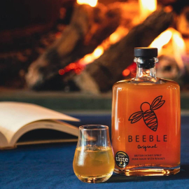 Image of the Beeble bottle set in a cosy interior, with a glass next to it, a book and a fireplace in the background. 