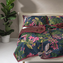 Kew Magnolia Blue Bedspread displayed on a bed with matching cushions. The deep blue quilt features a wonderful detailed print with rich fuchsia pink trumpets and forest green foliage with a contrasting block print design on the reverse. The reverse featu