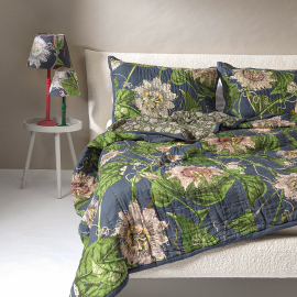 Kew Passion Flower Grey Bedspread displayed on a bed with matching cushions. The reverse features a cream floral design on a dark green background.