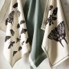 Lifestyle image of three tea towels hung, two featuring black and white artwork of insects and the other a duck egg blue with white striped lining.