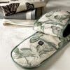 Lifestyle image of Kew Living Jewels Double Oven Glove with light grey background and illustrations of insects including moths and butterflies. Darker grey lining and dark grey inner. Kew Logo is featured in centre. Oven glove rested on surface with tea t