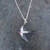 Image of the necklace featuring a swallow bird-shaped pendant in metal silver with navy accents. The necklace chain is in metal silver as the casting of the pendant and it lays on a marbled surface.