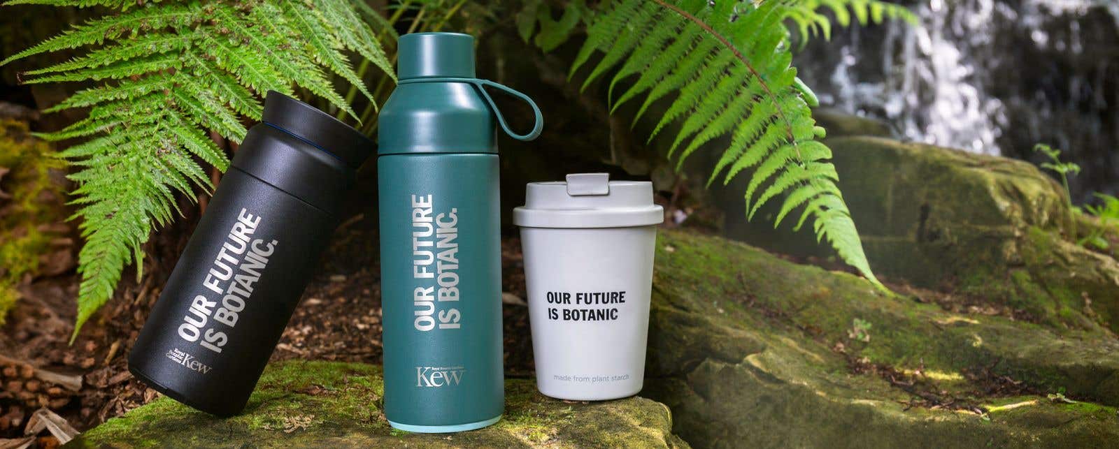 Two flasks and a reusable coffee cup resting on some rocks in front of a waterfall